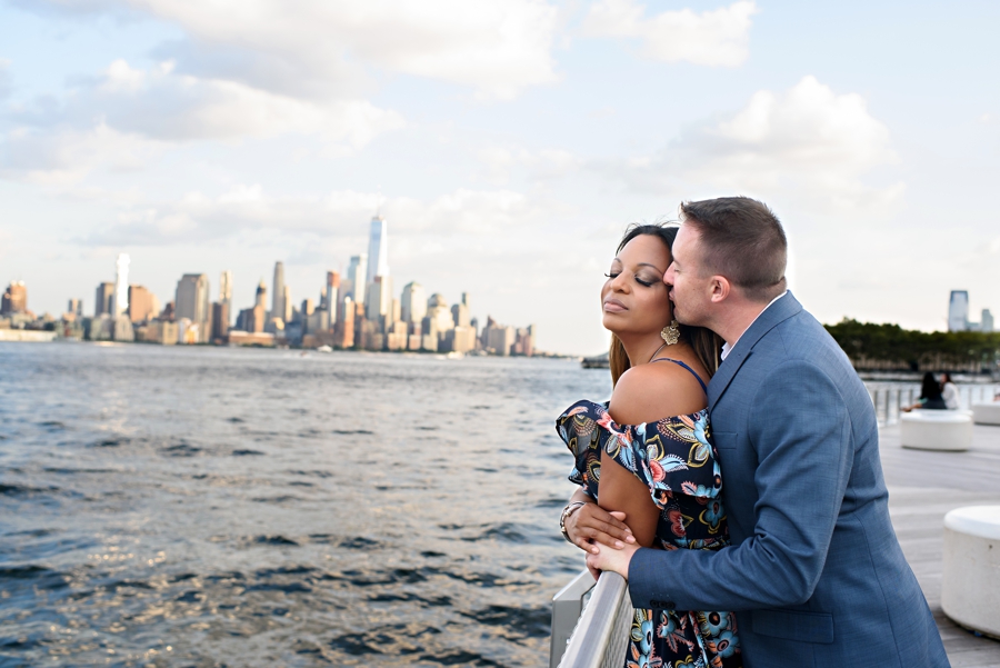 A man hugs is fiance while the wind blows her dress along the hudson river with manhattan skyline in the background.
